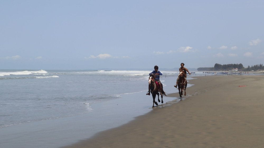 Two riders on horseback moving along a wide sandy beach with the sea behind them in bright daylight