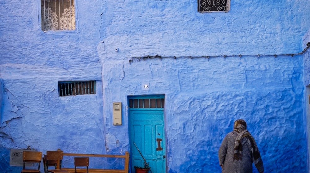 A person walking past a vivid blue-washed wall in a narrow street of Chefchaouen