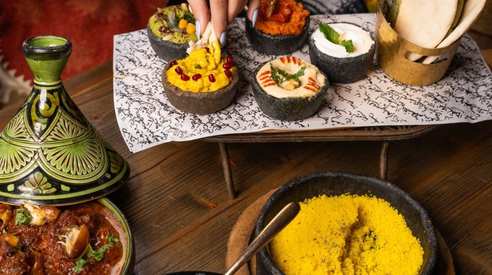 An overhead Moroccan dinner spread of beef tagine, couscous, bread and dips with a hand reaching in