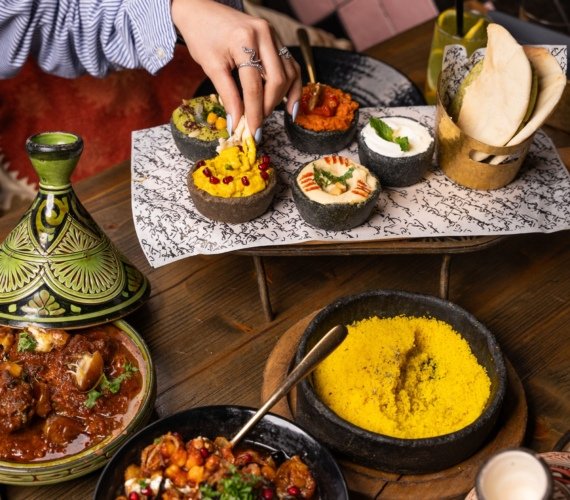 An overhead Moroccan dinner spread of beef tagine, couscous, bread and dips with a hand reaching in