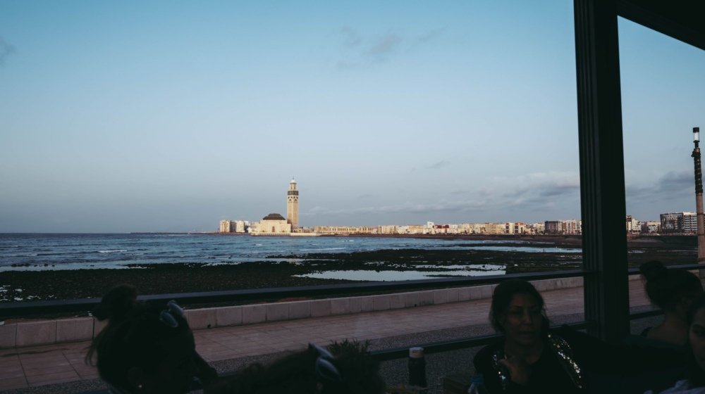 The Hassan II Mosque seen across the water from Casablanca's Atlantic seafront, with visitors seated at a coastal cafe terrace in the foreground