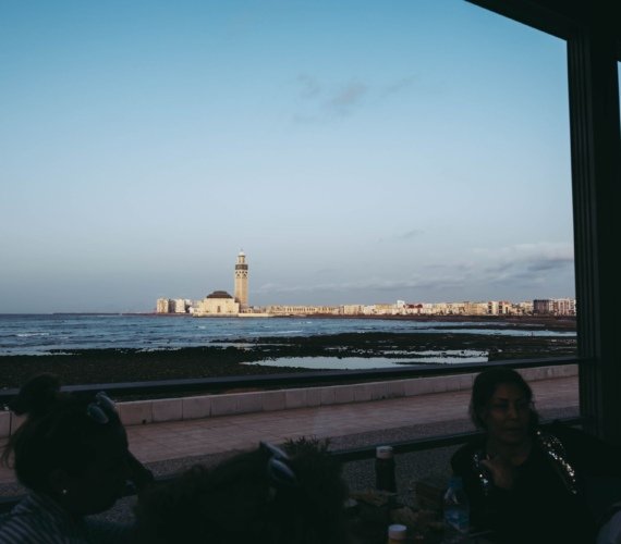 The Hassan II Mosque seen across the water from Casablanca's Atlantic seafront, with visitors seated at a coastal cafe terrace in the foreground