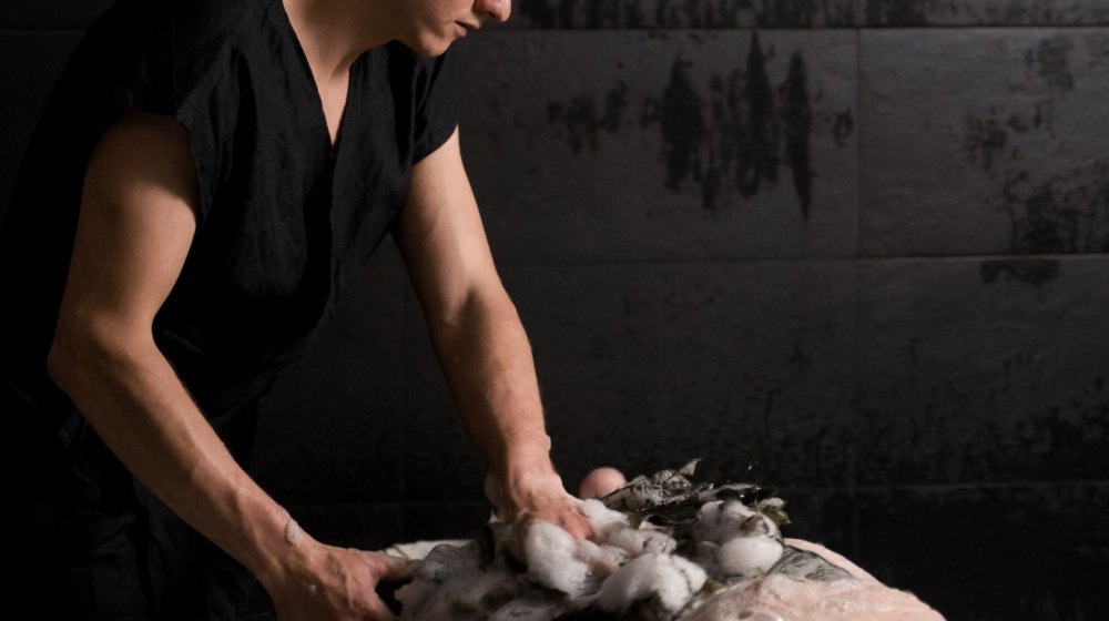 A therapist applying a soapy foam scrub to a client lying on a stone slab in a steam-filled hammam treatment room