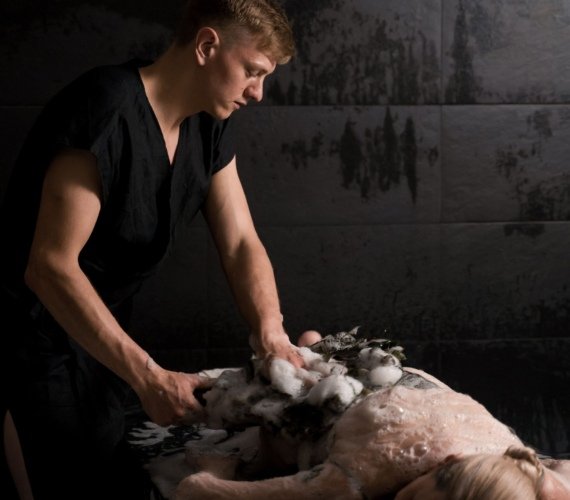 A therapist applying a soapy foam scrub to a client lying on a stone slab in a steam-filled hammam treatment room
