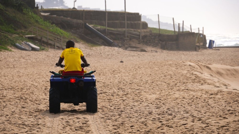 A rider driving a quad bike along the wet sand of an open beach with ocean surf breaking alongside