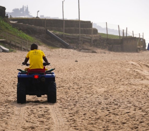 A rider driving a quad bike along the wet sand of an open beach with ocean surf breaking alongside