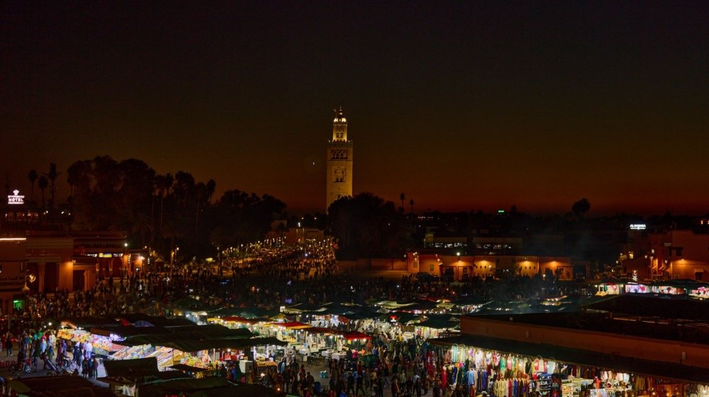 Jemaa el-Fna square in Marrakech at night, crowded with people among rows of illuminated food stalls, with the floodlit Koutoubia minaret behind.