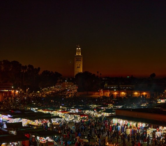 Jemaa el-Fna square in Marrakech at night, crowded with people among rows of illuminated food stalls, with the floodlit Koutoubia minaret behind.
