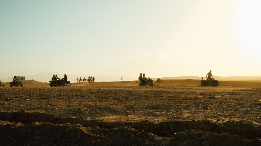 Quad bikers and a line of saddled camels together on the stony Agafay desert outside Marrakech at golden hour, with hazy hills behind.