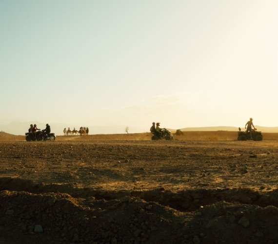 Quad bikers and a line of saddled camels together on the stony Agafay desert outside Marrakech at golden hour, with hazy hills behind.