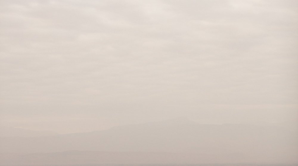 A green, yellow and red hot air balloon in flight over the red Marrakech plain, with a hazy mountain ridge along the horizon.