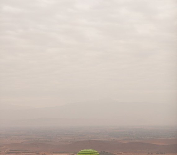 A green, yellow and red hot air balloon in flight over the red Marrakech plain, with a hazy mountain ridge along the horizon.