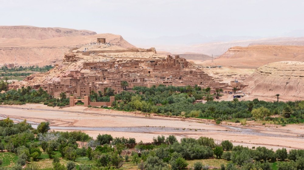 The fortified earthen ksar of Ait Ben Haddou near Ouarzazate rising above palm groves and a dry riverbed, with tiny visitors on the path to the summit watchtower.