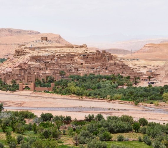 The fortified earthen ksar of Ait Ben Haddou near Ouarzazate rising above palm groves and a dry riverbed, with tiny visitors on the path to the summit watchtower.