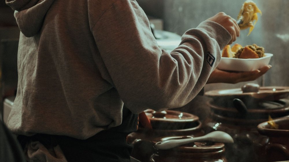 A cook in a patterned headwrap serving food from a row of steaming clay tagine pots in a warm Moroccan kitchen