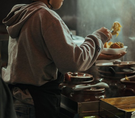 A cook in a patterned headwrap serving food from a row of steaming clay tagine pots in a warm Moroccan kitchen