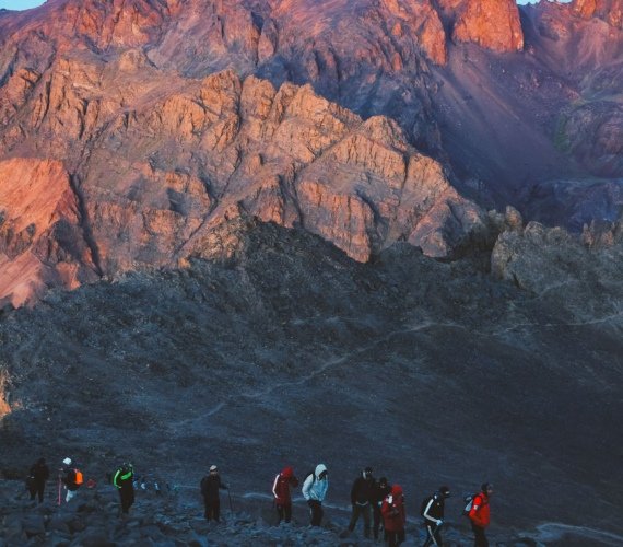 A line of trekkers crossing a rocky slope in the Moroccan mountains as first light hits the red rock face above