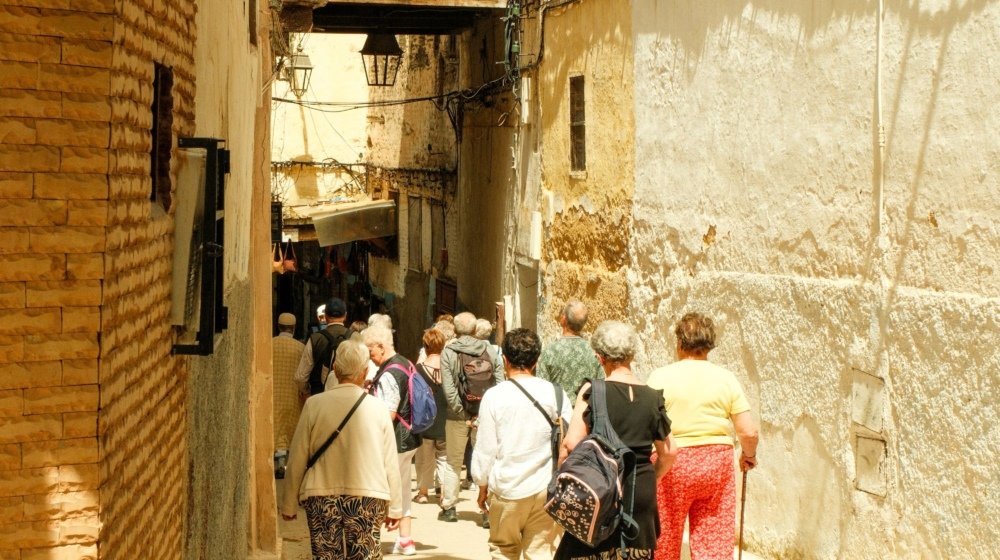 A group of visitors walking single file through a narrow sunlit lane in the Fez medina toward an archway and souk stalls
