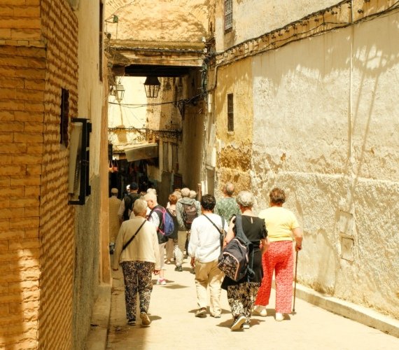 A group of visitors walking single file through a narrow sunlit lane in the Fez medina toward an archway and souk stalls