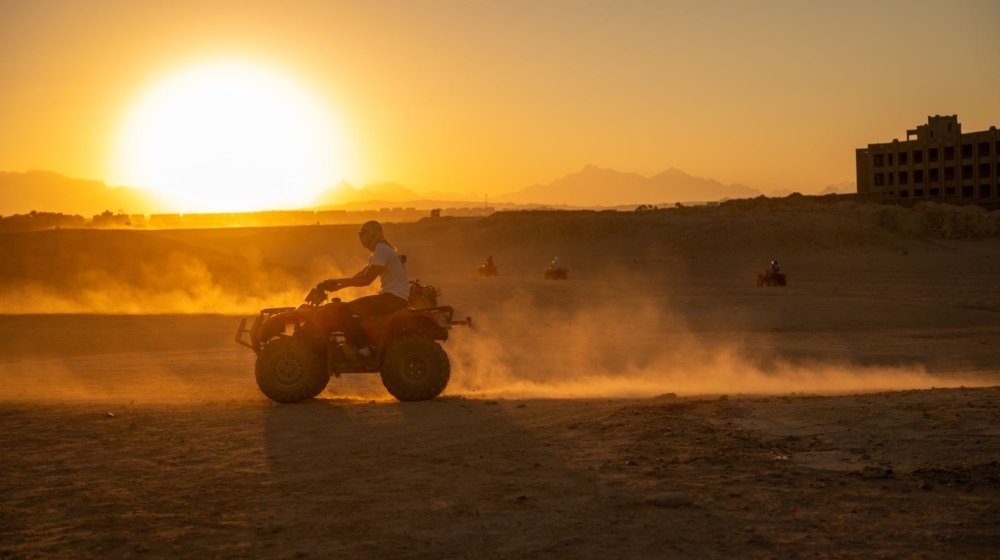 A group of quad bike riders crossing sand dunes in golden low sunlight with dust catching the light