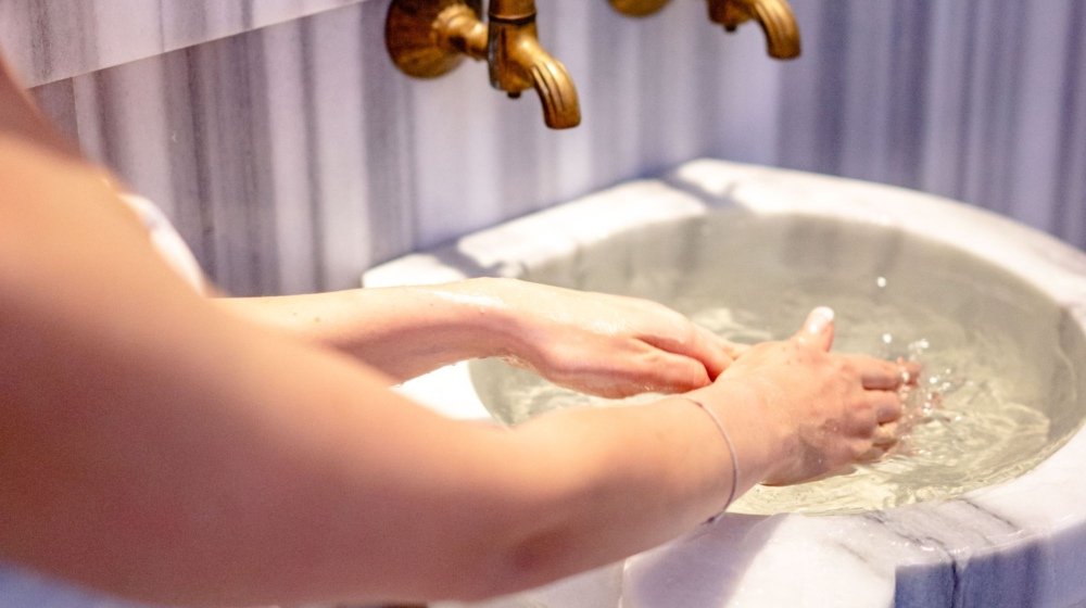 A woman rinsing her hands at an ornate marble basin with gold taps in a tiled bathhouse