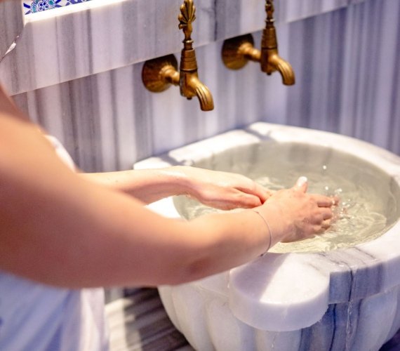 A woman rinsing her hands at an ornate marble basin with gold taps in a tiled bathhouse