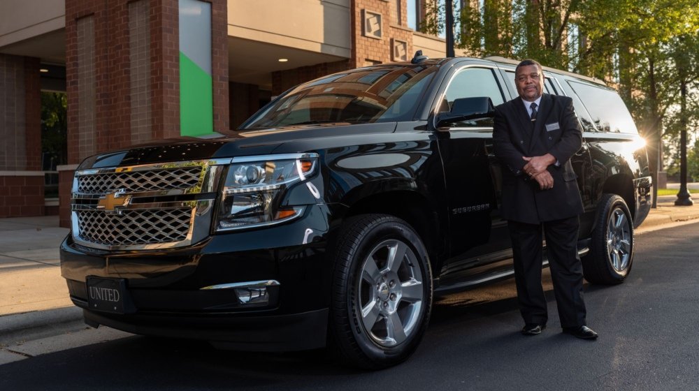 A uniformed chauffeur standing beside a black luxury SUV outside a modern city building