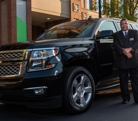 A uniformed chauffeur standing beside a black luxury SUV outside a modern city building